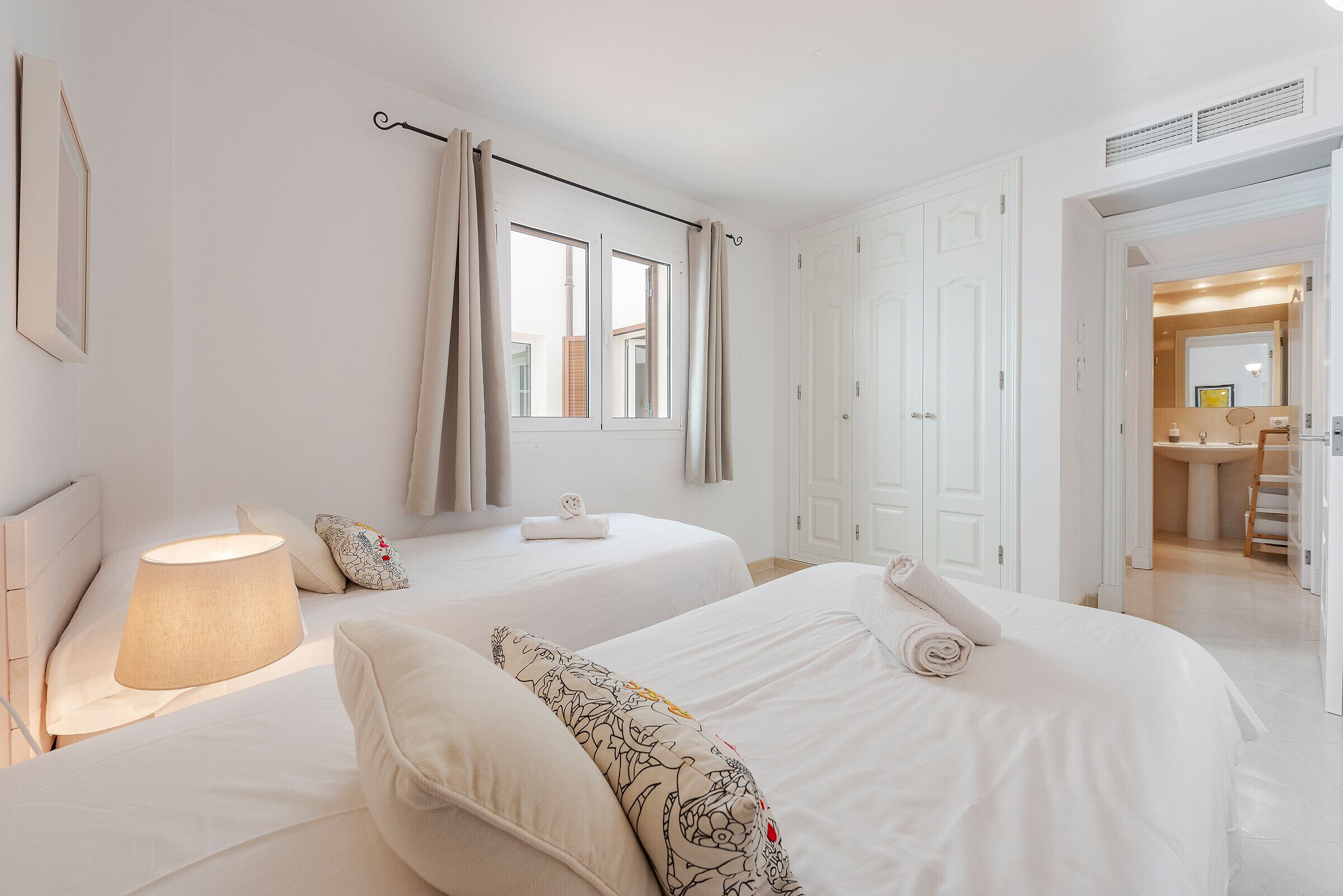 Une chambre d’hôtel avec deux lits, une fenêtre avec des rideaux, une lampe de chevet et une salle de bain visible à travers une porte ouverte.