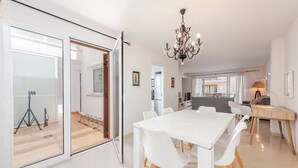 Et moderne spiseområde med et hvidt bord og stole, en lysekrone og udsigt gennem glasdøre til en balkon.