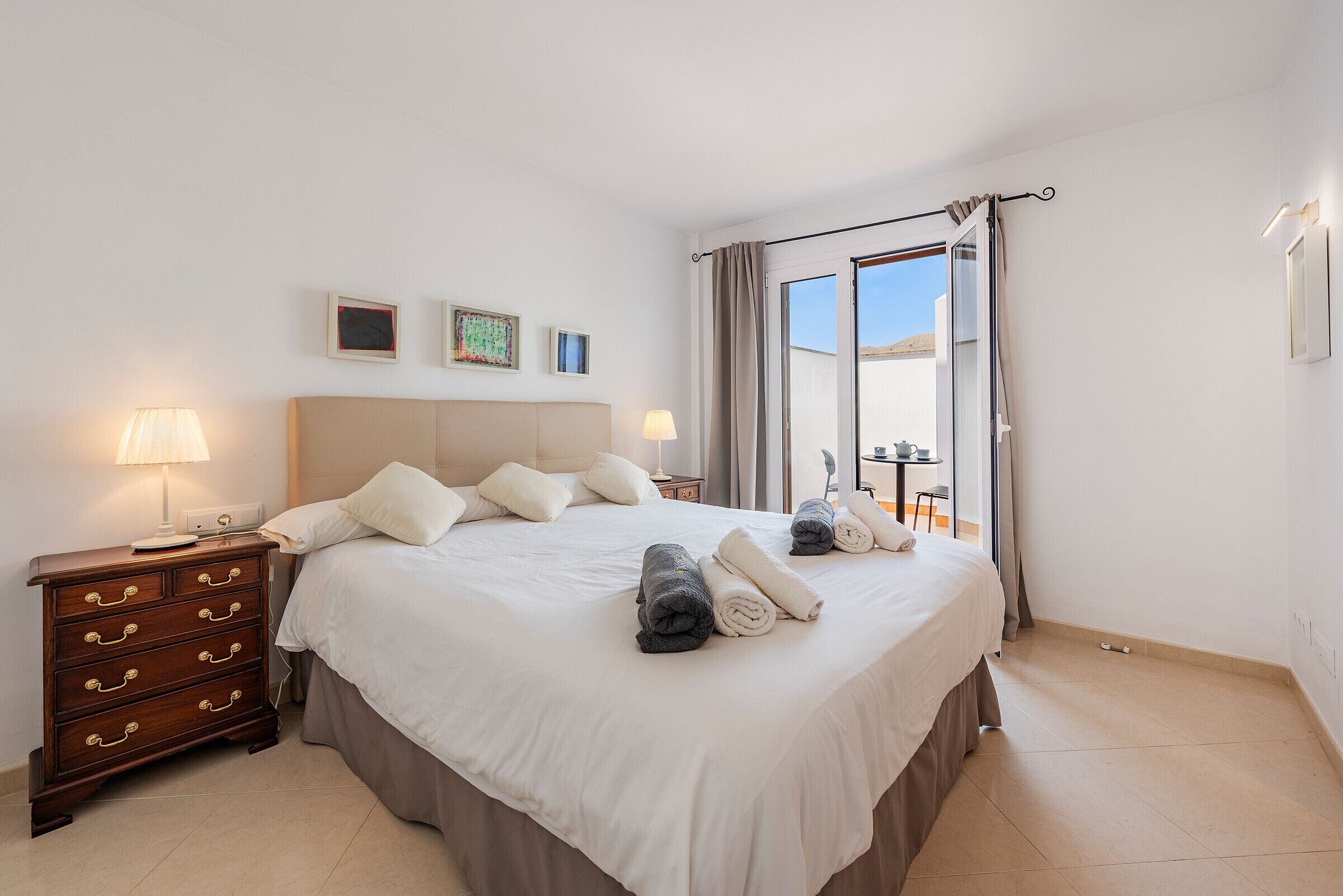 A neatly made bed with pillows and rolled towels, a bedside table with lamps, and a view of the outdoors through a sliding door.