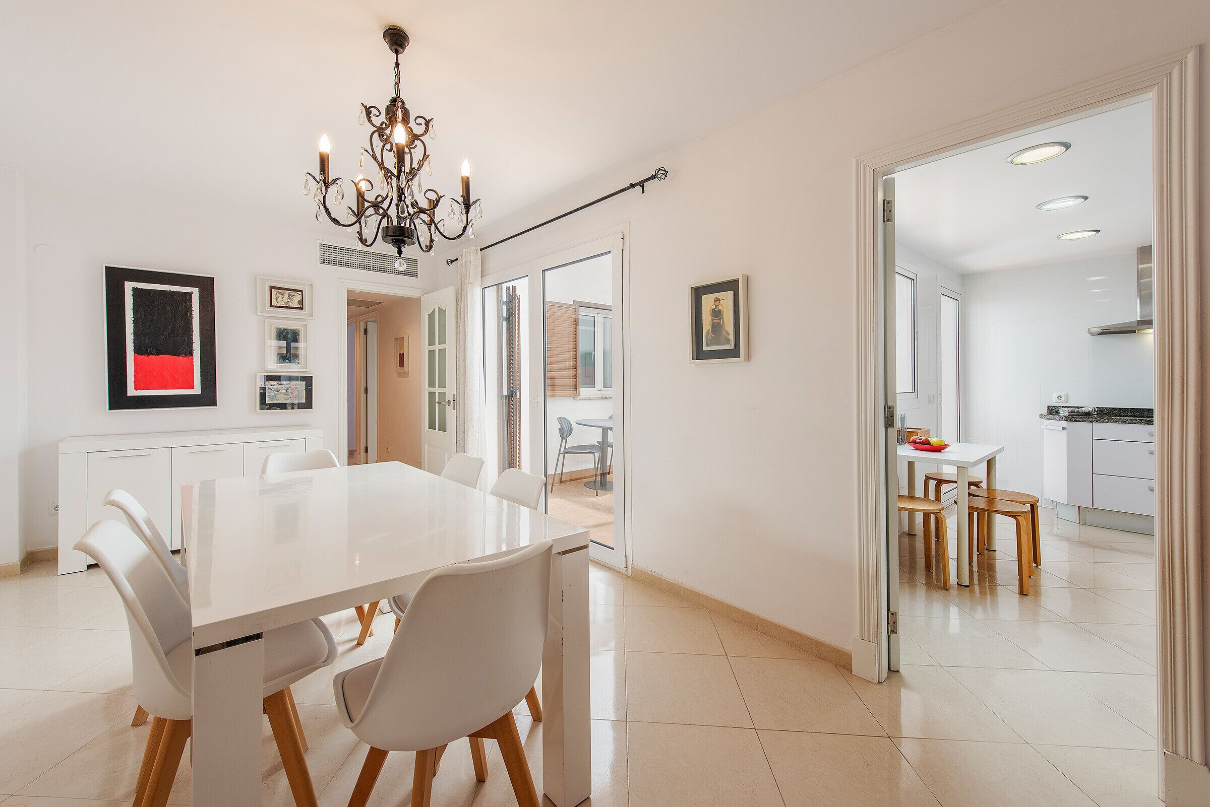 Un comedor moderno con una mesa y sillas blancas, una araña y vistas a la cocina.