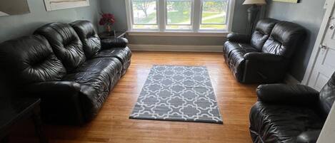 A living room with two black leather sofas, a patterned rug, wooden flooring, a ceiling fan, and a window with blinds.