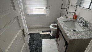 Een kleine badkamer met toilet, wastafel en raam met zonwering. Twee ingelijste foto’s hangen aan de muur en er is een douchestang.