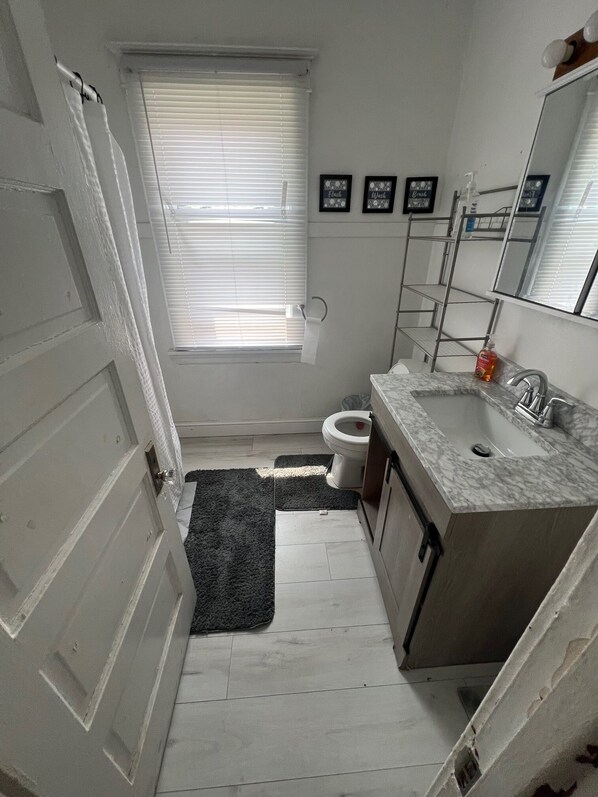 A small bathroom with a toilet, sink, and window with blinds. There are two framed pictures on the wall and a shower curtain rod.