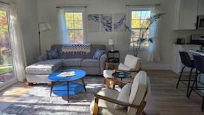 A modern living room with a sectional sofa, a blue coffee table, and a wooden chair. There is a kitchen area with a microwave and a toaster.