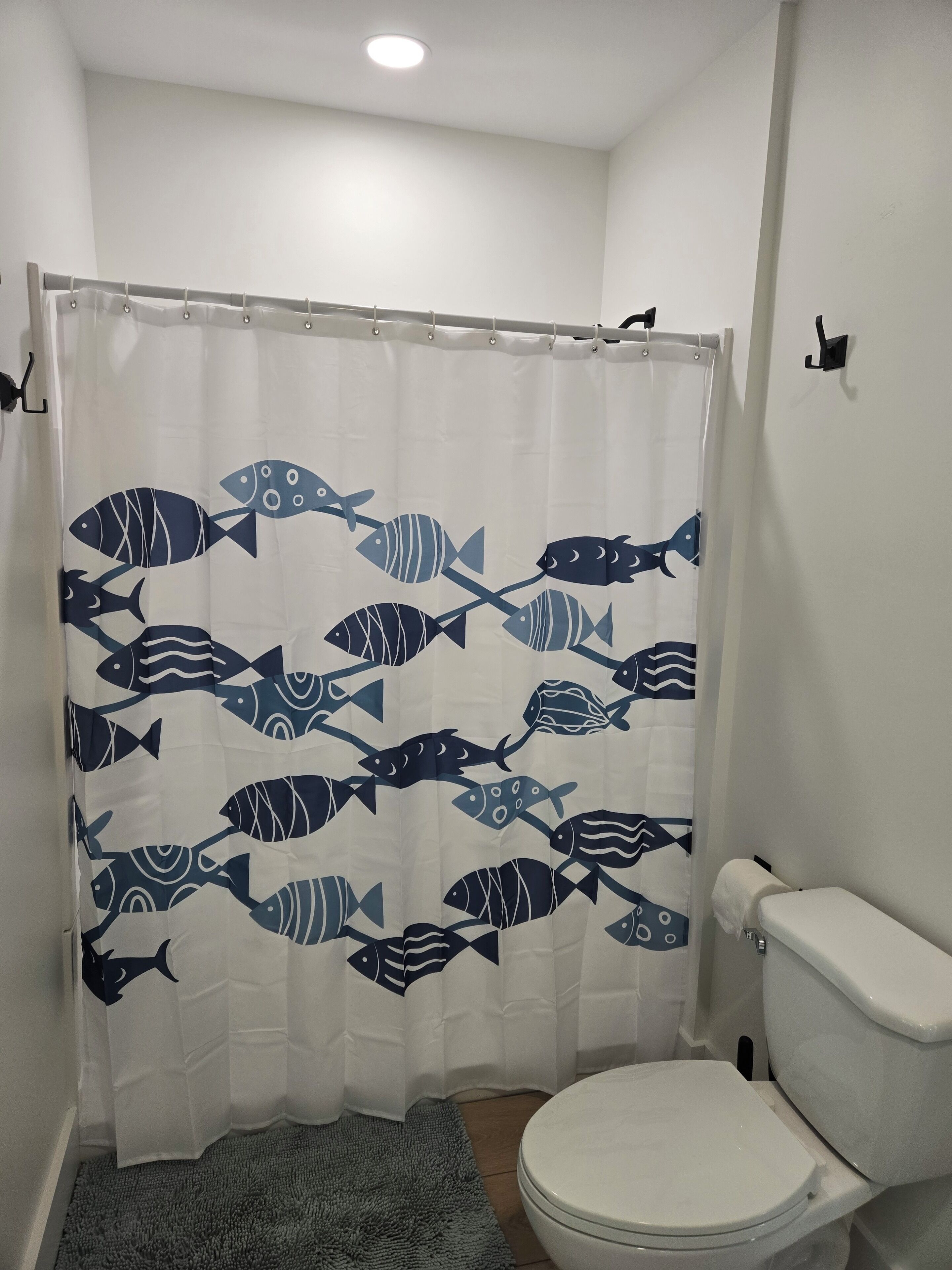 A bathroom with a white toilet, a shower curtain with a fish pattern, and a gray rug.