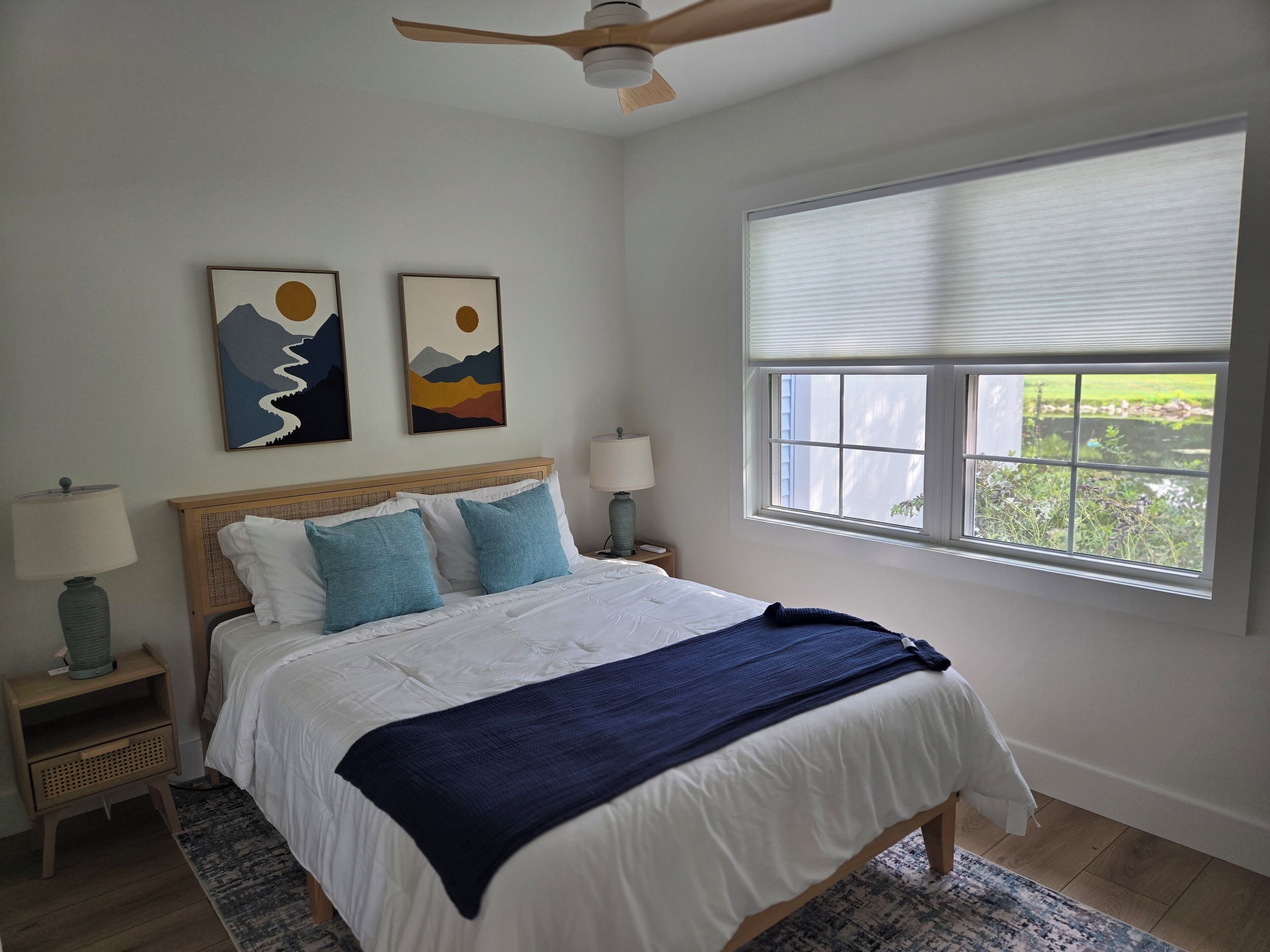 A bedroom with a bed, two wall-mounted paintings, a window with blinds, a ceiling fan, and a nightstand with a lamp.