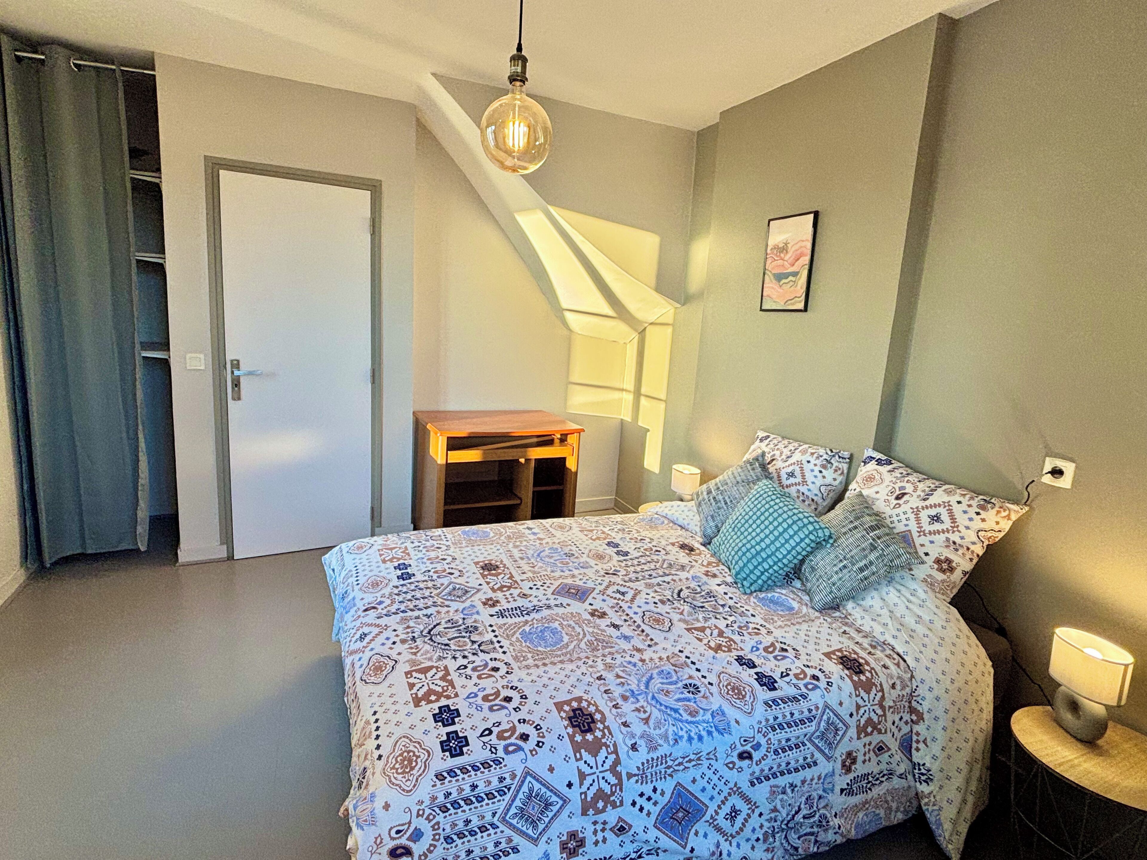 A bedroom with a bed, a nightstand, a desk, a chair, a door, and a window.
