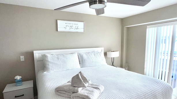 A neatly made bed with a folded towel, a bedside table with a tissue box, a ceiling fan, and a window with blinds. - Spacious Grand Haven Getaway (Grand Haven)