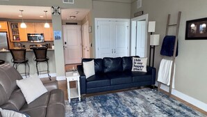 A spacious living area with a dark blue sofa, a brown leather chair, and a blue patterned rug. A white door and a white railing are visible in the background. - Spacious Grand Haven Getaway (Grand Haven)