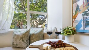 A cozy seating area with a wooden board displaying cheese, grapes, and crackers, accompanied by two glasses of red wine, set against a backdrop of a large window with a view of trees and a building. - Stylish Studio with Rooftop Harbour Views (Darling Point)