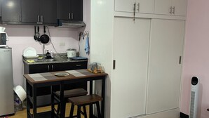 A compact kitchen with dark cabinets, a white fridge, a small dining table with stools, and a white wardrobe with a mirror. - Welcome to Solstay, a get go place to stay Cozy 1-bedroom condo near Alabang (Muntinlupa)