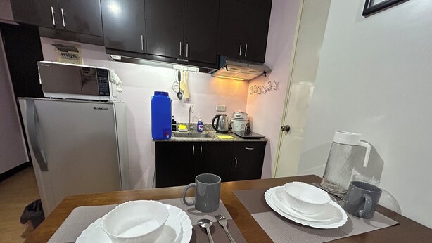 A compact kitchen with dark cabinets, a refrigerator, a sink, and a coffee maker. There is a dining table set with plates, a cup, and cutlery. - Welcome to Solstay, a get go place to stay Cozy 1-bedroom condo near Alabang (Muntinlupa)