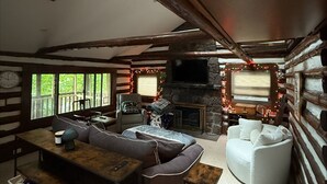 A cozy log cabin interior with a stone fireplace, wooden beams, a large window, and a comfortable seating area.