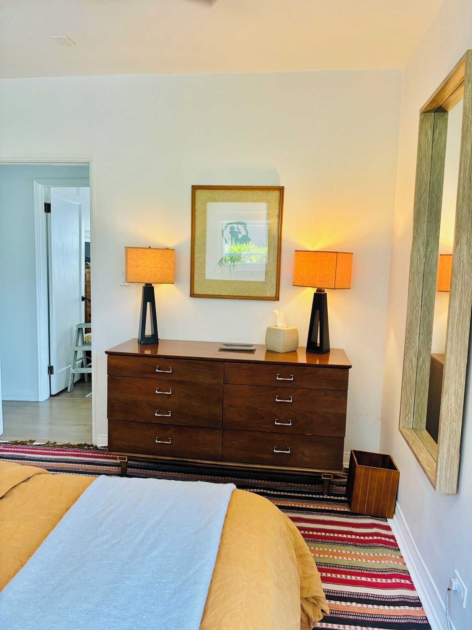 A bedroom with a bed, a dresser, two wall-mounted lamps, a framed picture, and a mirror.
