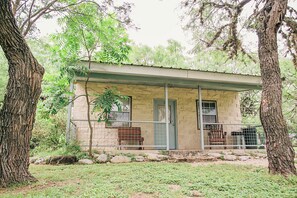 A small stone building with a porch, surrounded by trees and rocks. - Nice Little Cabin in the Woods, Frio River Frontage - #3 (Concan)