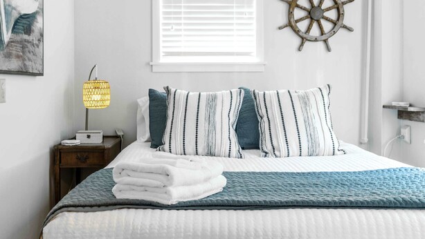 A neatly made bed with a striped pillow set, a ship's wheel decoration on the wall, and a bedside lamp. - Williams Bay Lakefront Retreat - Walk the Lakepath (Williams Bay)
