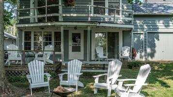 A two-story house with a porch, multiple Adirondack chairs, and a wheelbarrow with firewood.