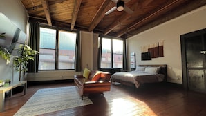 Ein geräumiges Zimmer mit Holzbalkendecke, einem Bett, einer Couch, einem Fernseher und großen Fenstern.