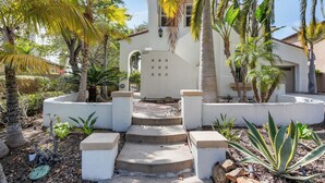 A white building with a number '6213' on the door, surrounded by palm trees and a concrete planter with steps leading to the entrance.