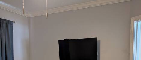 Un dormitorio con cama, mesita de noche con lámpara, televisor montado en la pared y ventana con cortinas.