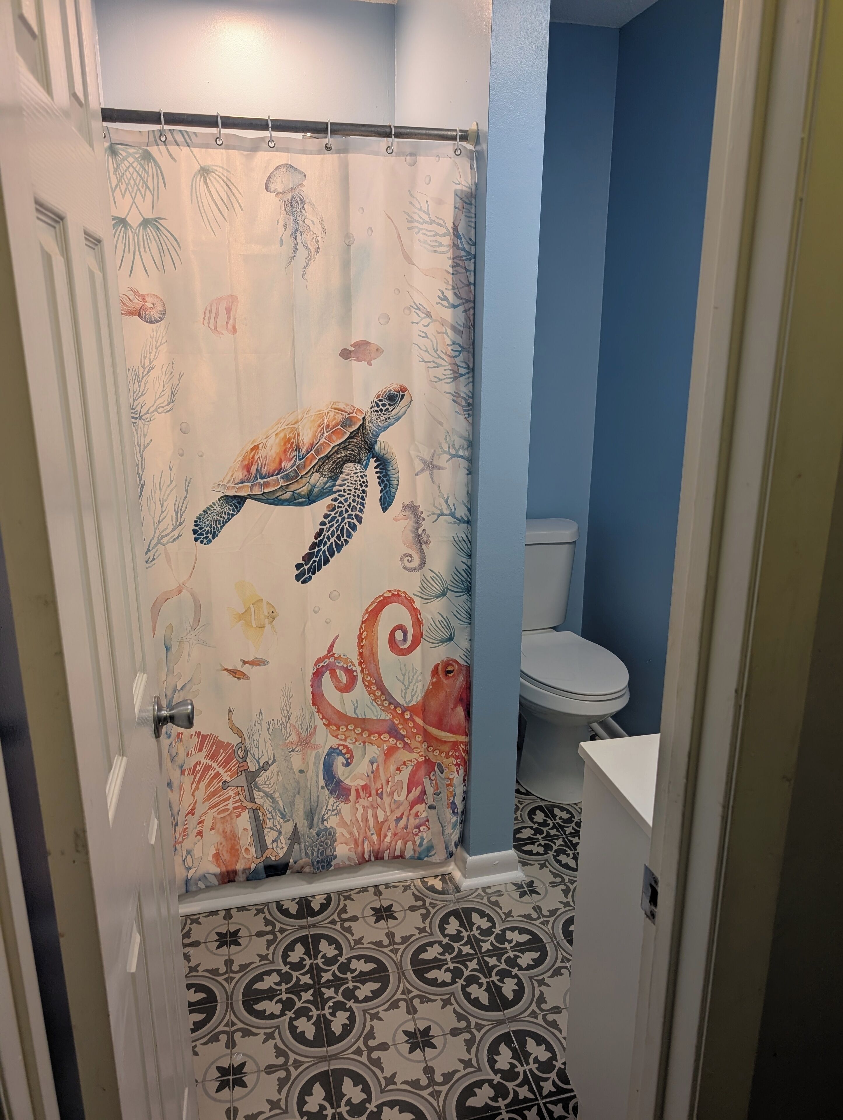 Ein Badezimmer mit Duschvorhang, der Meeresschildkröten und Meereslebewesen zeigt, einer Toilette und fliesenbelagtem Boden mit Muster.