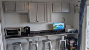 Eine kleine Küche mit Einbauofen, Kaffeemaschine und einem Computer auf der Arbeitsfläche. Drei Metallhocker und eine Schachtel Ocean Blue sind ebenfalls vorhanden.