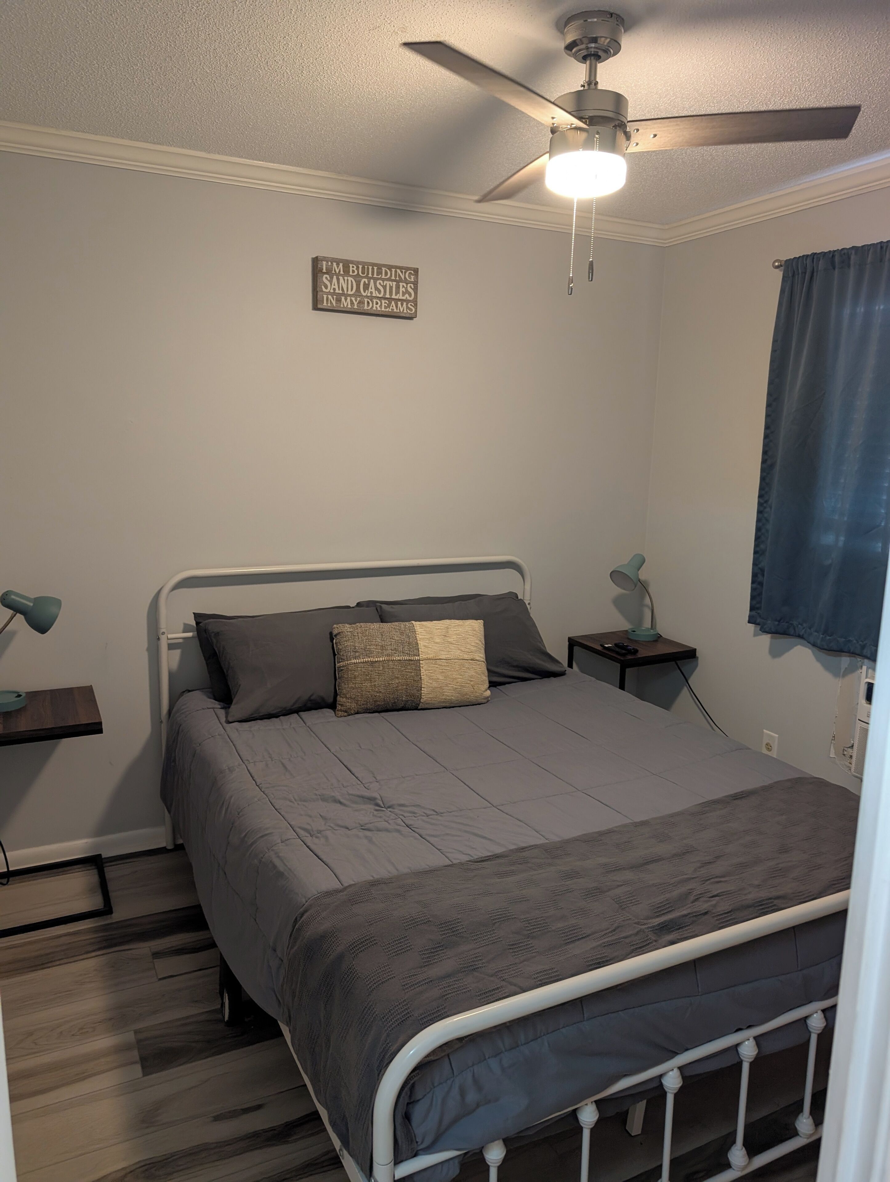 Una camera da letto con un letto, una ventola a soffitto, un comodino con una lampada e un cartello a parete.