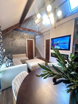 A modern living room with a wooden dining table, white sofa, and a large TV screen displaying Penarth Pier. - Luxury Penarth Loft - Near Cardiff; Great for Entertainment and Sporting Events (Penarth)
