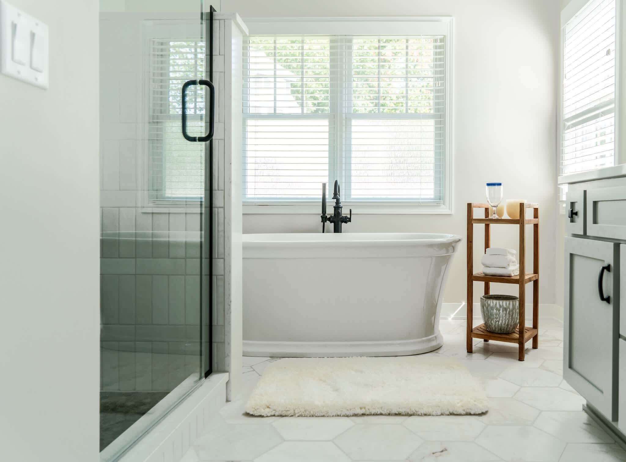 A bathroom with a glass shower enclosure, a white bathtub, a wooden shelf with toiletries, and a window with blinds.