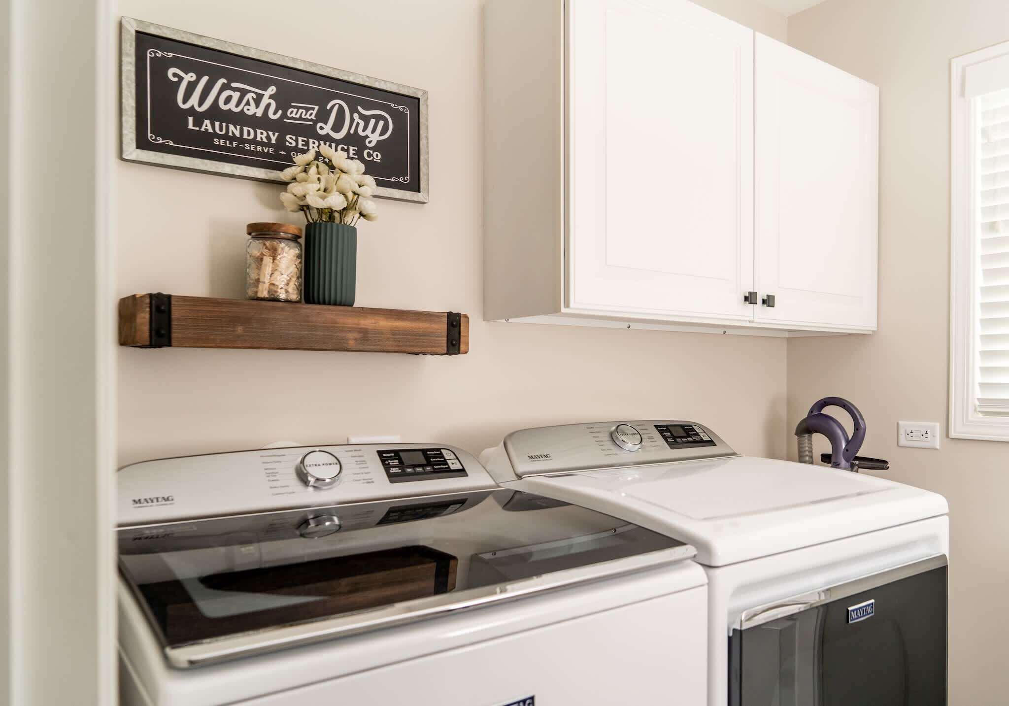 Un área de lavandería con dos lavadoras y secadoras Maytag, un estante con un florero y frascos, y un letrero que dice 'Wash and Dry Laundry Service Co.'