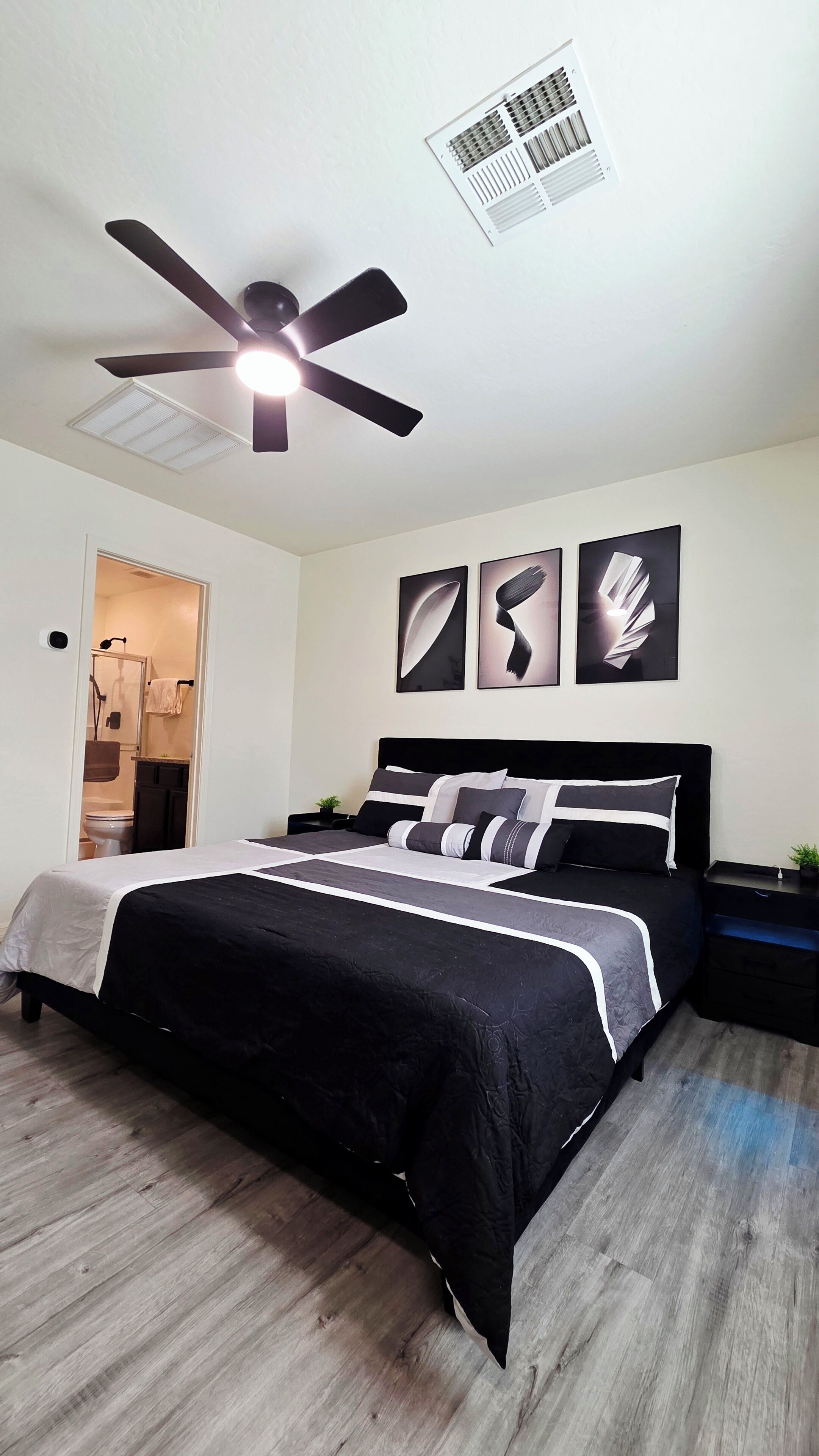 A bedroom with a bed, a ceiling fan, a wall-mounted air vent, and three abstract wall art pieces.