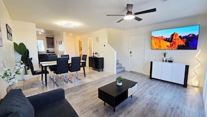 Una sala de estar moderna con un televisor de pantalla plana, un ventilador de techo, un comedor con sillas y una mesa, y una cocina al fondo.