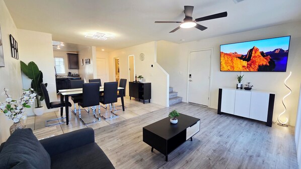 A modern living room with a flat-screen TV, a ceiling fan, a dining area with chairs and a table, and a kitchen in the background.
