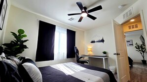 A hotel room with a bed, a desk, a chair, a ceiling fan, and a window with curtains.