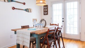 A dining area with a wooden table, chairs, a woven pendant light, and a 'PRAY FOR SURF' sign on the wall.
