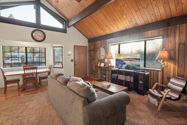 Un salón acogedor con techo de madera, un ventanal con vistas, una zona de comedor con mesa y sillas, y una estantería.