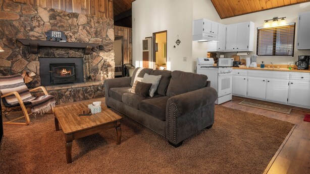 A cozy living room with a stone fireplace, a sofa, a wooden coffee table, and a kitchen area with white cabinets and appliances.