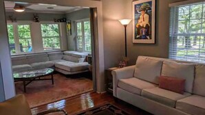 A living room with a white sofa, a glass coffee table, and a patterned rug. There are two windows with blinds, a lamp, and a framed picture on the wall.