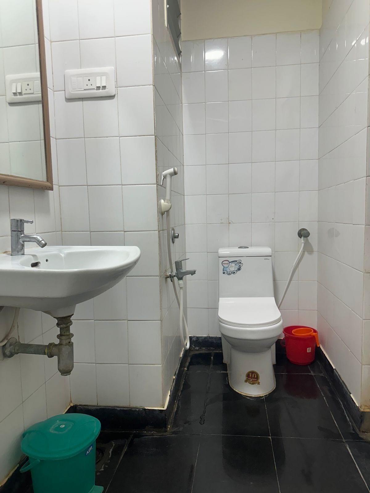 A small, tiled bathroom with a white sink, toilet, and shower. There is a green trash bin and a red bucket on the floor.
