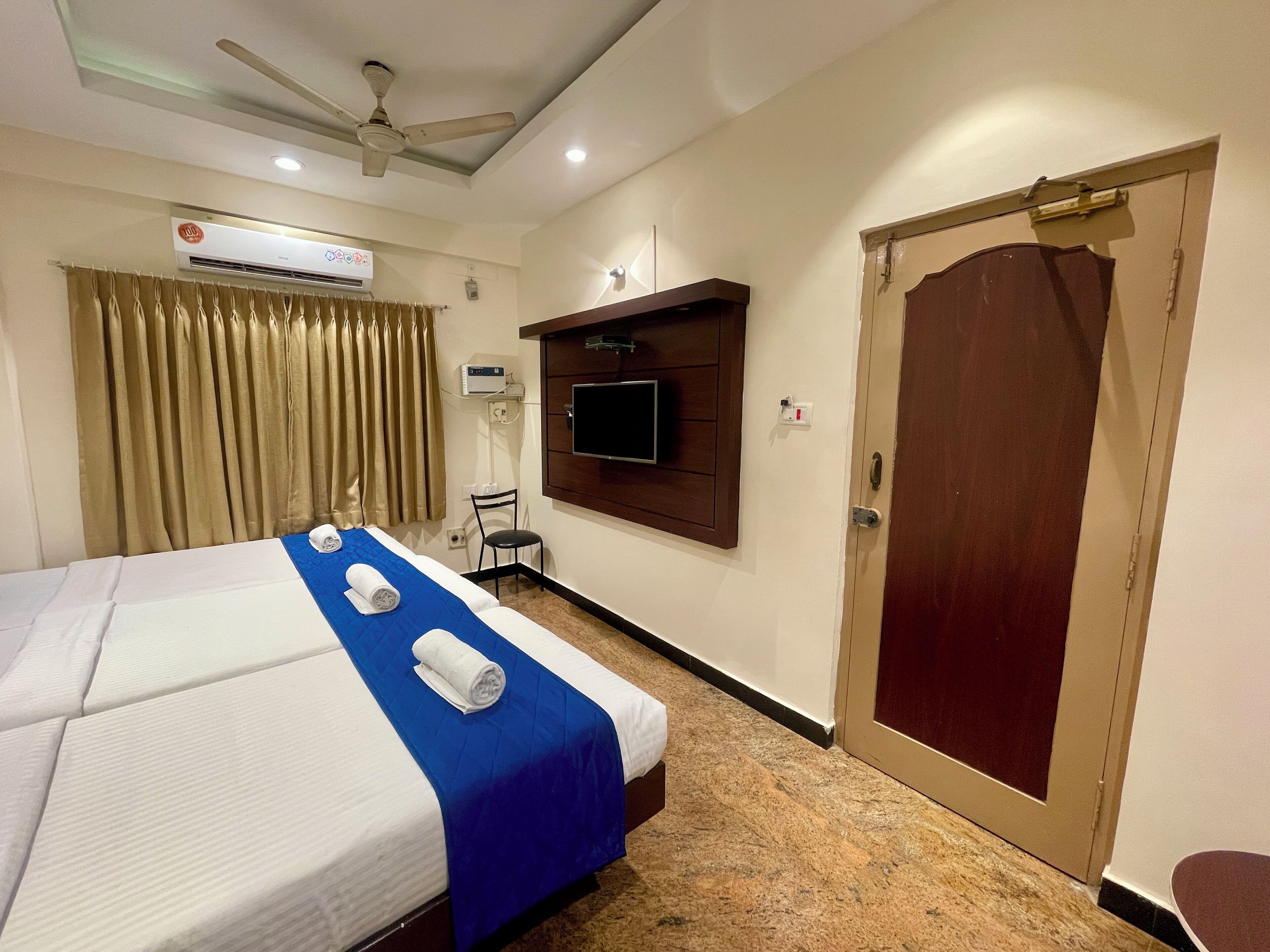 Een hotelkamer met een bed, een plafondventilator, een airconditioningsysteem, een aan de muur bevestigde televisie en een houten deur.