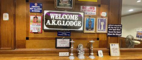 A reception desk with a welcome sign, framed pictures, and various notices including no smoking and room charges.
