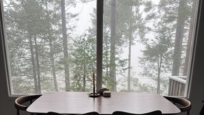 A dining area with a table and chairs, a candle, and a view of a forest through a large window. - The Crescent Moon Cabin with Lakeviews & Wood burning Fireplace (Crestline)