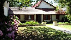 Une maison de plain-pied avec un toit en tuiles, un porche à l’avant et un jardin bien entretenu agrémenté de fleurs roses.