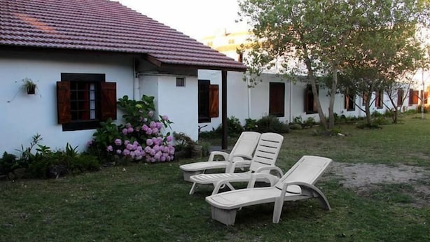 Un bâtiment blanc avec un toit recouvert de tuiles, un jardin parsemé de fleurs roses et deux fauteuils de jardin blancs.