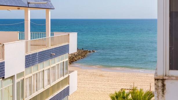 Un edificio frente a la playa con un balcón que da al mar, una playa de arena y un litoral rocoso.