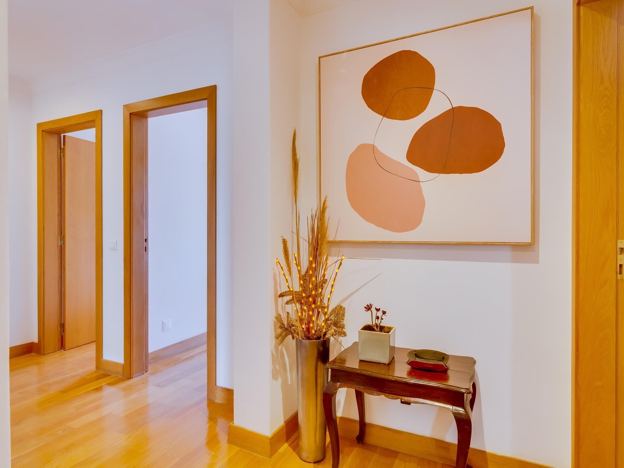 A hallway with wooden flooring, a vase with dried plants, a framed abstract painting, and a small wooden table with a red object on it.