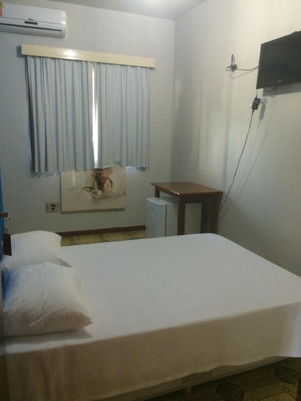A hotel room with a bed, a small table, a TV mounted on the wall, and an air conditioning unit.