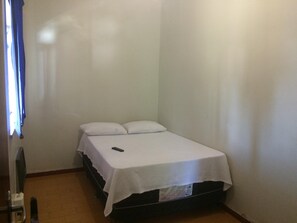 Une petite chambre avec un lit simple, recouverte d’une courtepointe blanche, avec deux oreillers et une télécommande posée sur le lit.