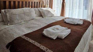 A bedroom with a large bed, a headboard, a painting on the wall, and a window with curtains. - Apart Raices Patagonicas (Dina Huapi)
