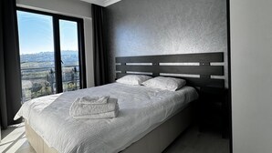 Une chambre moderne avec un grand lit, une table de chevet et une vue sur le paysage urbain, visible à travers une grande fenêtre.